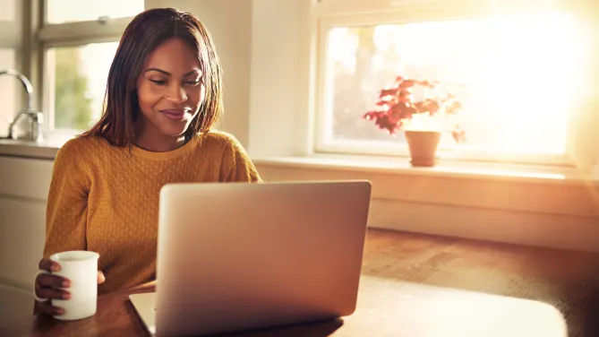 Woman smiling looking at computer with cup in hand.
