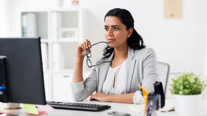 Woman thinking at desk with glasses to her chin.