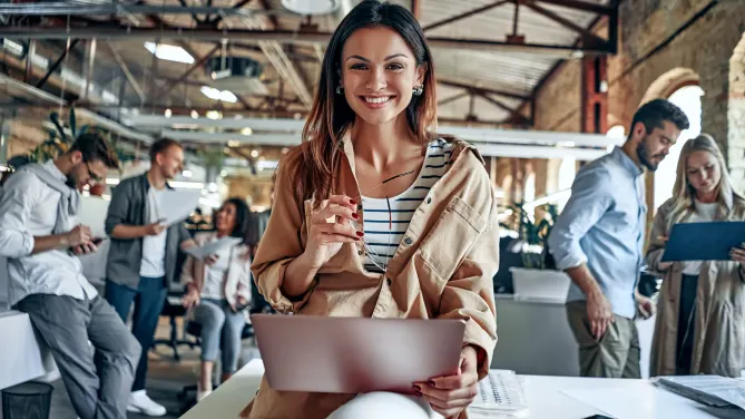 Woman in an office smiling at the camera with a laptop on her lap. People working in the background.