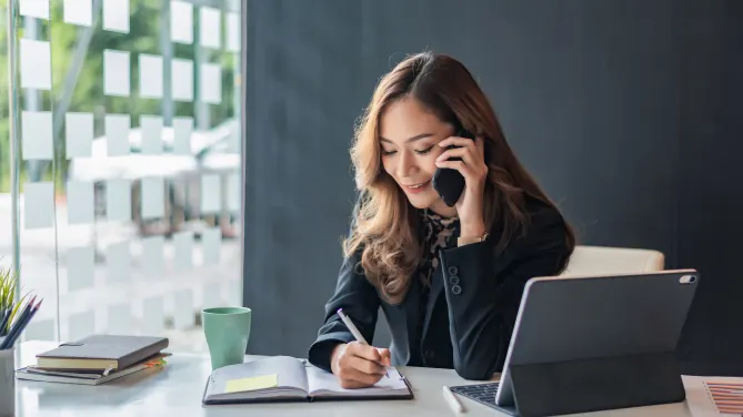 Woman talking on the cell phone while writing in a notebook.