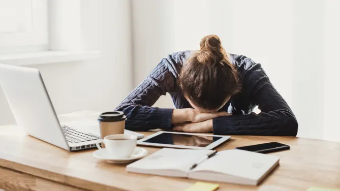 Woman with face down and arms crossed on desk. Coffee, notebook, laptop and tablet in front of her.