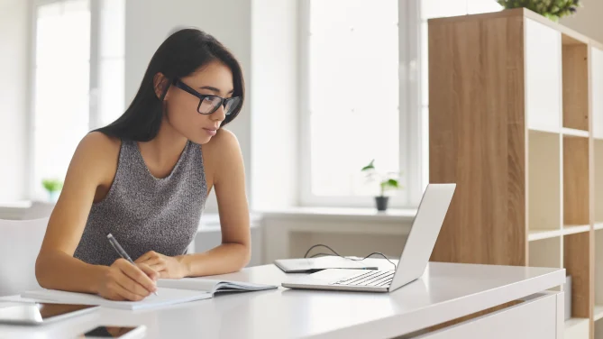 Woman taking notes in notebook with laptop in front of her.