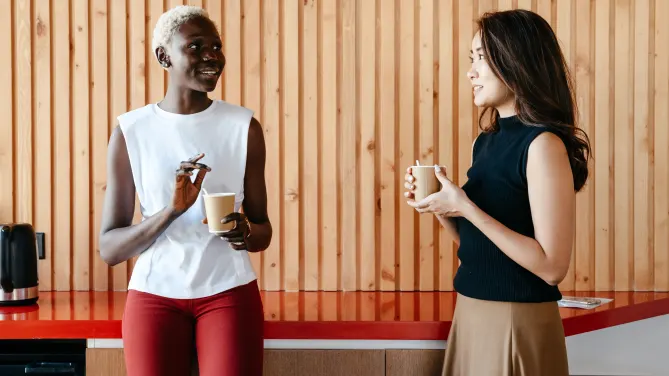 two women holding paper coffee cups chatting by a wood panel wall