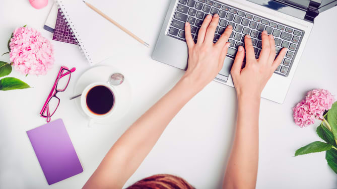 woman typing on desk with flowers, notepad and glasses