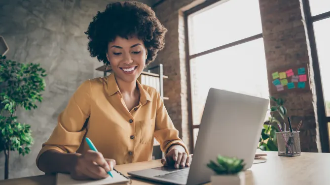 Woman smiling at notebook on desk to her right with a pen in hand. Her left hand sits on her laptop trackpad. 