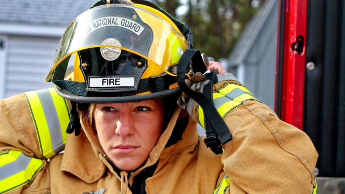 Woman military firefighter lowers yellow helmet onto her face while assessing her situation.