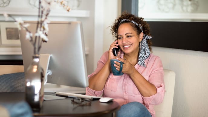 Woman sitting at desk with a desktop computer. She talking on the phone and smiling with a mug in hand.