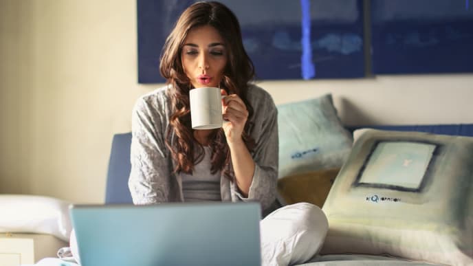 A woman sits in bed sipping coffee while working on a laptop