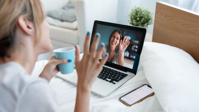 Woman waving at computer screen, which shows another woman waving back.