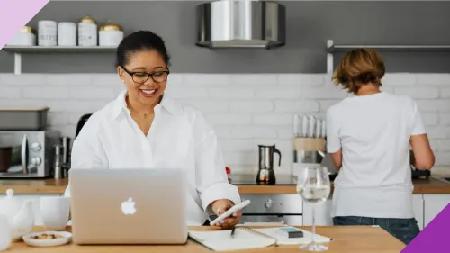 Two women in the kitchen of their house, one of them is managing finances on phone and computer, illustrating how family budget apps can make your life easier