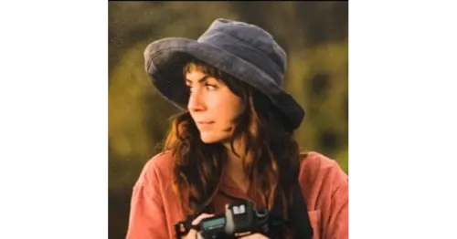 Jessi is standing in a field, wearing a hat and a corduroy shirt, holding a camera and looking to the right. 