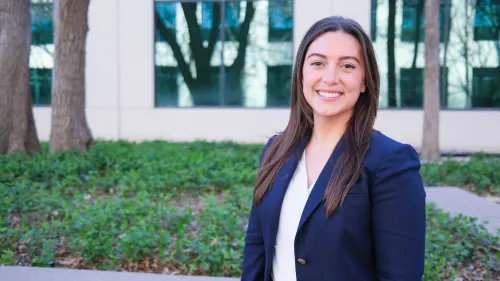 Francesca is standing outside an office building in a blue blazer, smiling. 