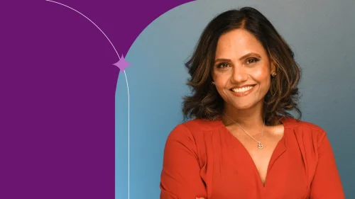 A photo of Priyanka Lingwal. She has shoulder length brown hair and is smiling at the camera. She is wearing a dark orange shirt standing in front of a dusty blue background.