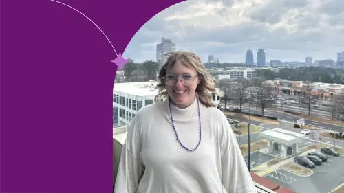 Ali standing in front of a window, there's a town behind her. she's similing and wearing white blose