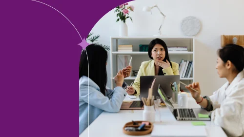 Three coworkers having a focused team meeting around a desk, discussing ideas with notebooks in a bright, modern office setting