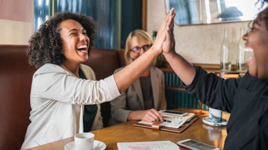 Two women of color hi-fiving across a table 