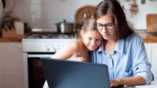 Woman looking at laptop with young girl resting on her shoulder, looking on.
