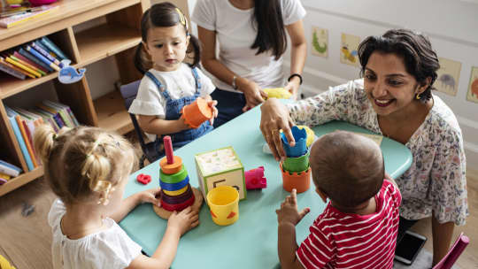 preschool teacher sitting at table playing with toddlers and blocks