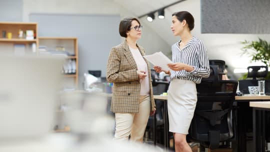Side view portrait of two contemporary businesswomen discussing work plans while standing in office, copy space