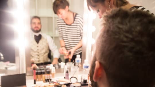 An actor sitting in a dressing room getting his makeup done by a makeup artist